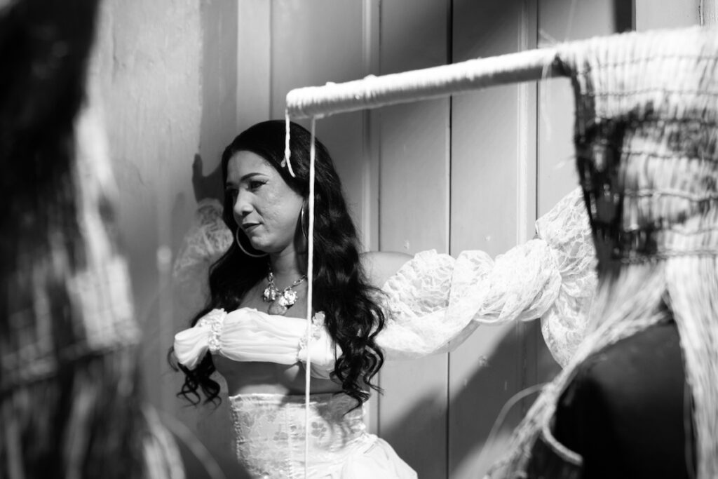 Uma mulher em um vestido branco com mangas de renda e longos cabelos escuros está entre duas peças de tecido em uma sala mal iluminada, olhando para o lado. A imagem é em preto e branco.