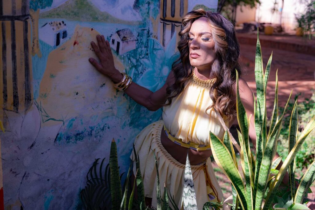 Uma mulher em uma roupa amarela está ao lado de um mural colorido, tocando a parede com uma das mãos. Ela está cercada por plantas verdes e banhada pela luz do sol, com os olhos fechados e uma expressão de calma no rosto.