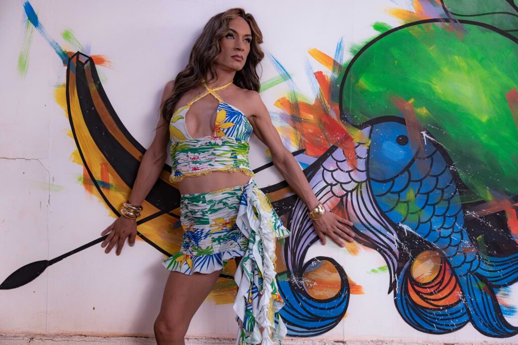 Uma pessoa em uma roupa tropical colorida posa confiante contra uma parede pintada com um mural de um peixe azul estilizado e formas abstratas vibrantes.