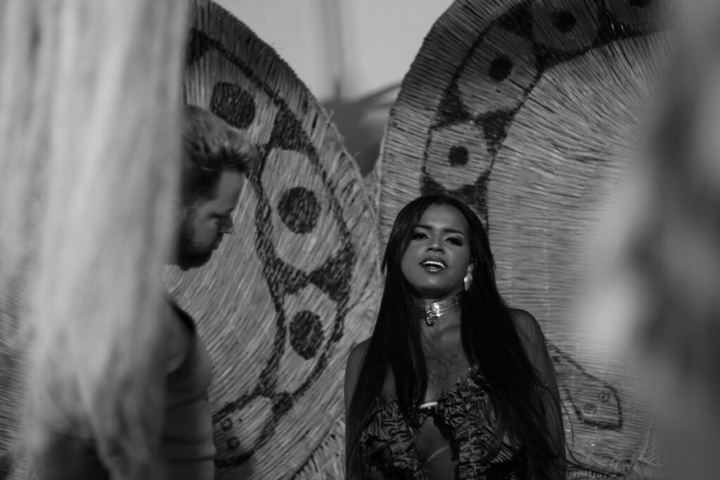 Uma mulher com longos cabelos escuros e um colar de gargantilha está em frente a grandes decorações circulares de tecido, enquanto um homem olha para ela. A imagem é em preto e branco.