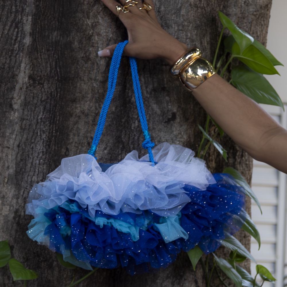 Uma mão usando joias de ouro segura uma bolsa de mão azul e branca com babados e uma alça de corda azul, posicionada em frente a um tronco de árvore cercado por folhas verdes.