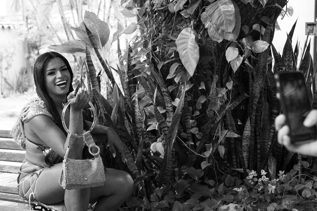 Uma mulher sentada em um banco ao ar livre, sorrindo e segurando uma bolsa, enquanto alguém tira uma foto dela com um telefone. Plantas exuberantes preenchem o plano de fundo. A imagem é em preto e branco.