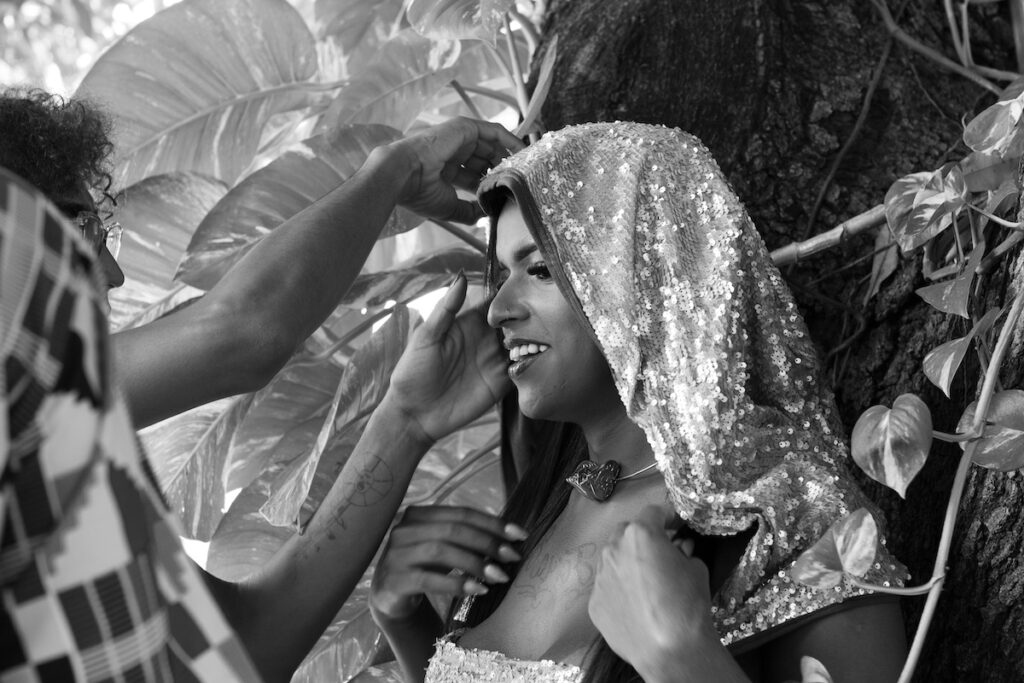 Uma pessoa usando um capuz brilhante com lantejoulas sorri enquanto outra pessoa ajusta sua roupa. Elas estão ao ar livre, perto de folhas grandes e de um tronco de árvore. A imagem é em preto e branco.