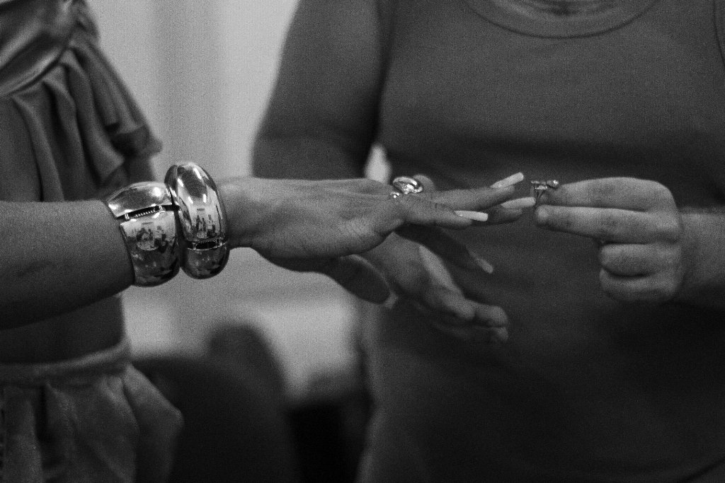 Close-up em preto e branco de uma pessoa colocando um anel no dedo de outra. A mão do destinatário está adornada com grandes pulseiras metálicas e ambas as figuras estão vestindo roupas sem mangas. O ambiente parece íntimo.