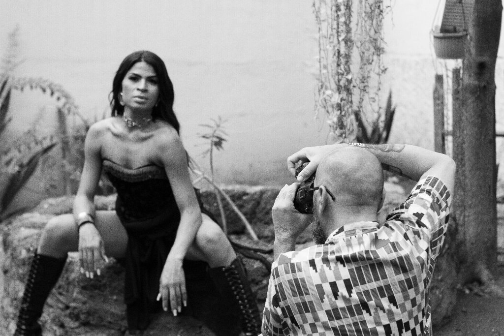 Uma pessoa posa confiante ao ar livre, sentada em uma pedra com as pernas afastadas, enquanto outra pessoa com uma camisa estampada a fotografa. A cena é em preto e branco com plantas ao fundo.