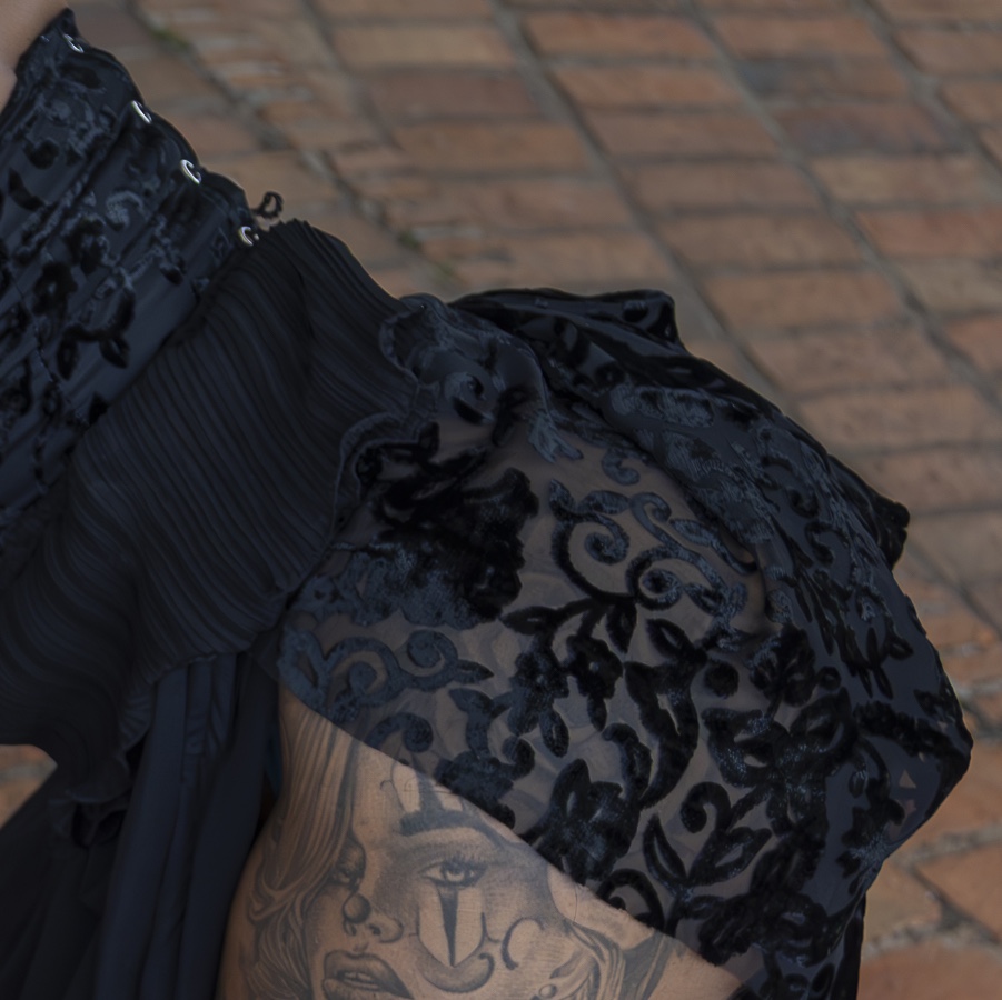 Um close-up de uma pessoa vestindo uma roupa preta, transparente, com estampa floral, com rendas e pregas visíveis, mostrando uma tatuagem detalhada de um rosto de mulher na parte superior do braço. O fundo é uma superfície de tijolos.