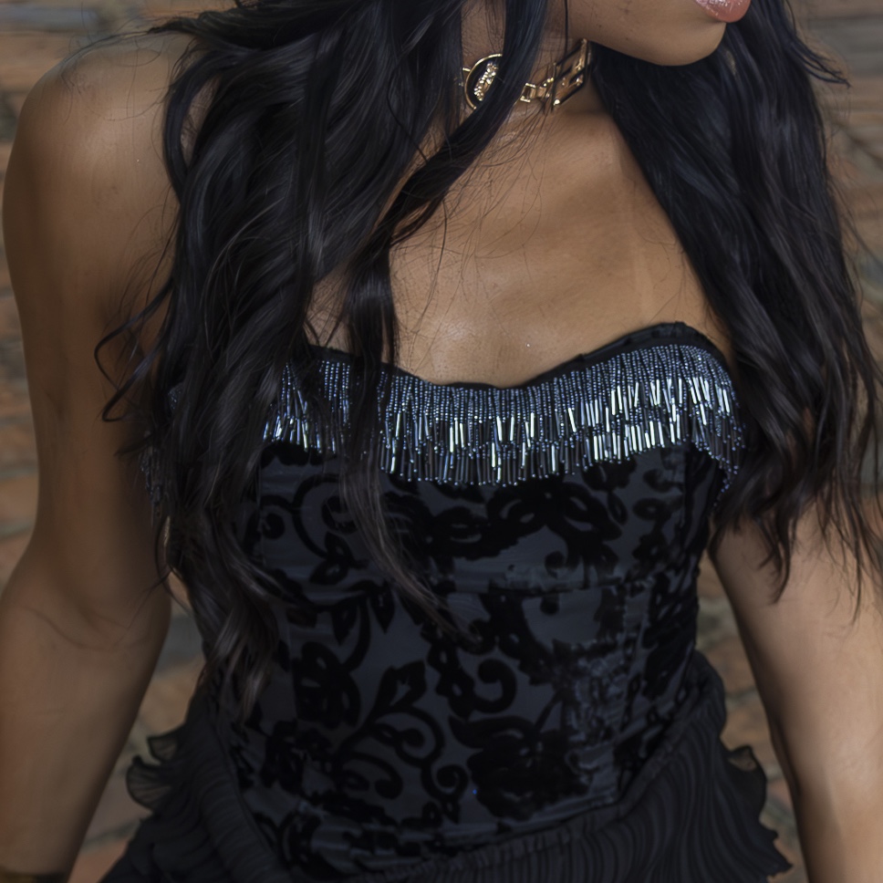 Uma mulher de cabelos escuros longos e ondulados usa um vestido de veludo preto sem alças com miçangas azuis e prateadas na parte superior e um colar de gargantilha dourado. O fundo apresenta uma parede de tijolos embaçada.