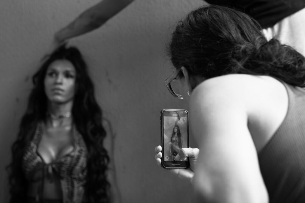 Uma pessoa com cabelo comprido posa contra uma parede enquanto outra pessoa tira uma foto dela com um smartphone. Uma terceira mão ajusta o cabelo da modelo. A cena é em preto e branco.