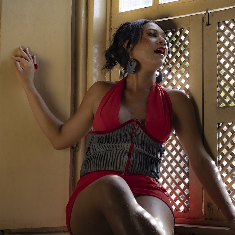 Uma mulher em um vestido vermelho com um espartilho listrado está sentada em uma janela iluminada pelo sol, olhando para cima com a boca ligeiramente aberta e uma mão apoiada na parede. Ela usa brincos de argola grandes e parece relaxada.