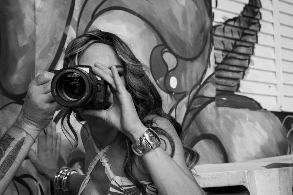 Uma pessoa de cabelos longos segura uma câmera no rosto, tirando uma foto. Ela está usando um relógio e uma pulseira, com um mural pintado na parede atrás dela. A imagem é em preto e branco.