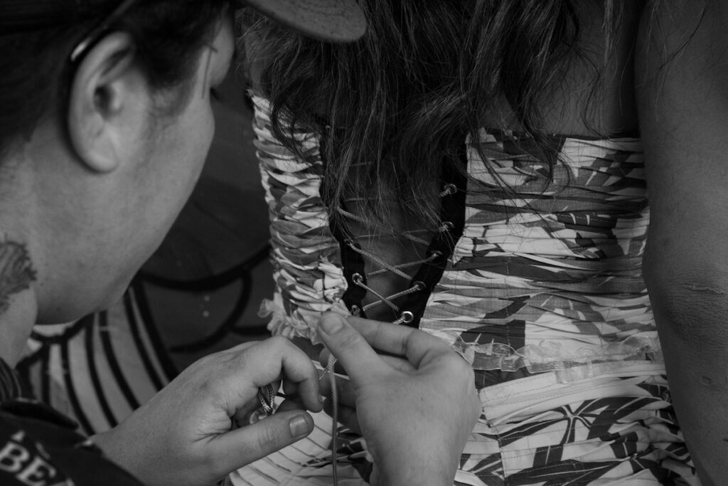 Uma pessoa está amarrando a parte de trás do vestido estampado de outra pessoa. A imagem é em preto e branco, com foco nas mãos que amarram os cadarços e nos detalhes intrincados do vestido.