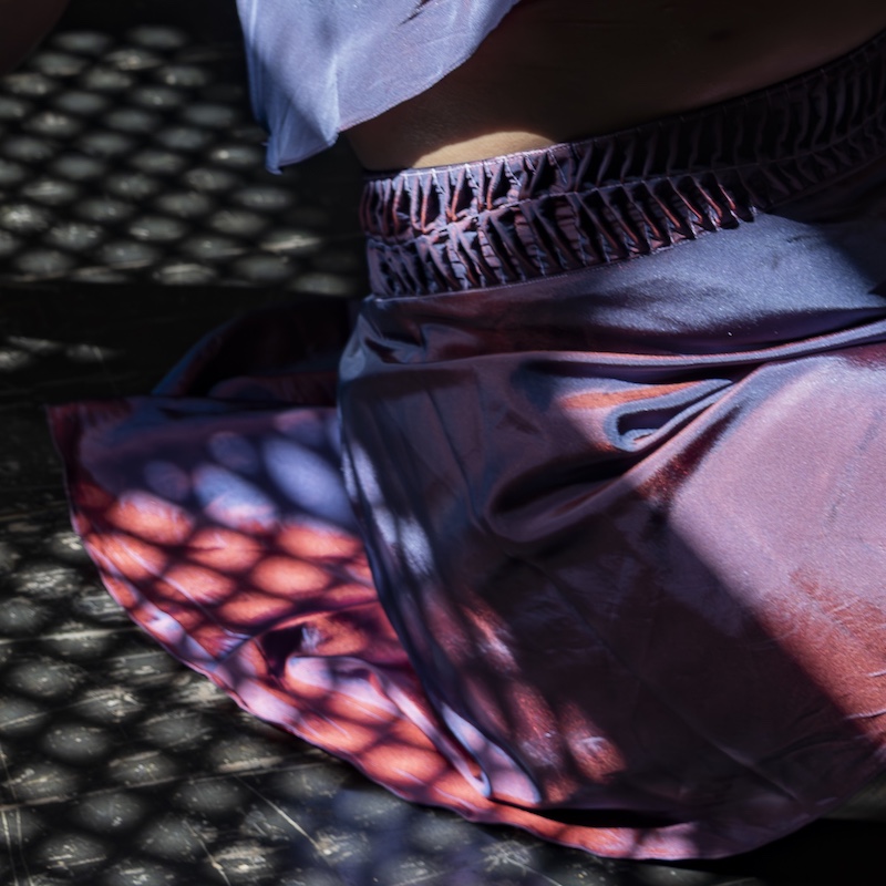 Uma pessoa vestindo uma saia brilhante, roxa e rosa senta-se no chão sob a luz do sol filtrada por uma superfície com padrão de grade, criando um desenho de sombra em suas roupas e pele.