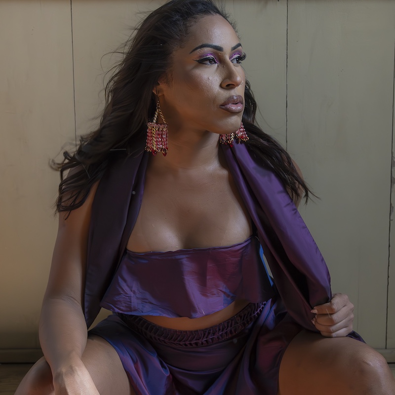 Uma mulher com longos cabelos escuros veste um traje de cetim roxo com brincos que combinam. Ela se senta contra uma parede de madeira clara, olhando para os lados com uma expressão confiante.