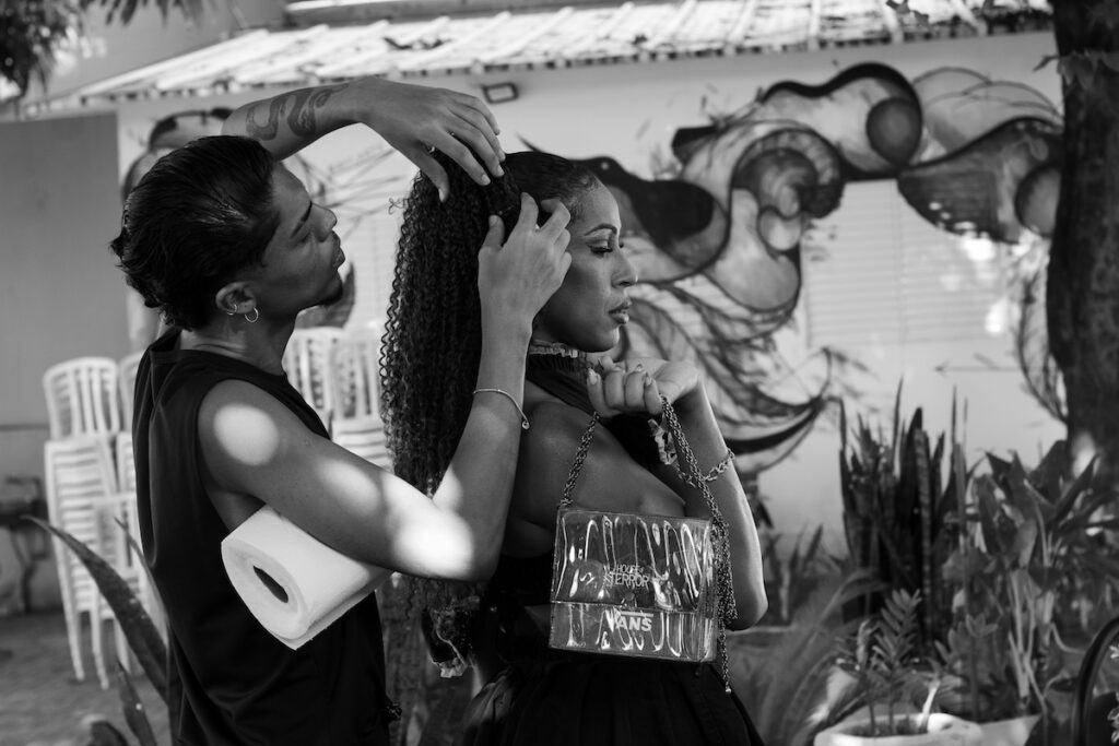 Uma pessoa penteia o cabelo cacheado de outra pessoa ao ar livre. A pessoa que está sendo penteada segura uma bolsa transparente e está em frente a um mural e a plantas. A cena é em preto e branco.