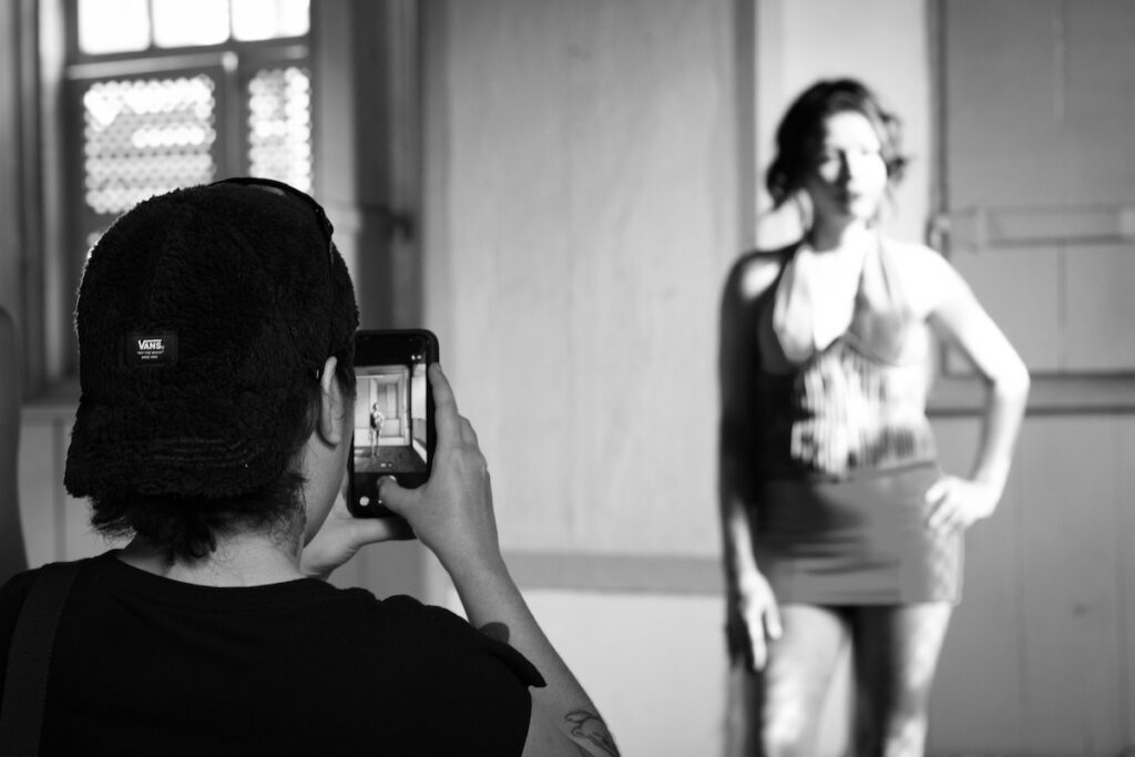 Uma pessoa usando um boné tira uma foto com seu telefone de outra pessoa de vestido, posando em frente a uma parede bem iluminada em uma sala com janelas. A imagem é em preto e branco.