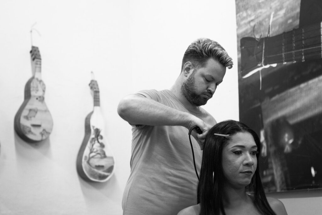 Um cabeleireiro usa uma chapinha para alisar o cabelo de uma mulher em um salão de beleza. Dois violões decorativos estão pendurados na parede ao fundo. A imagem é em preto e branco.