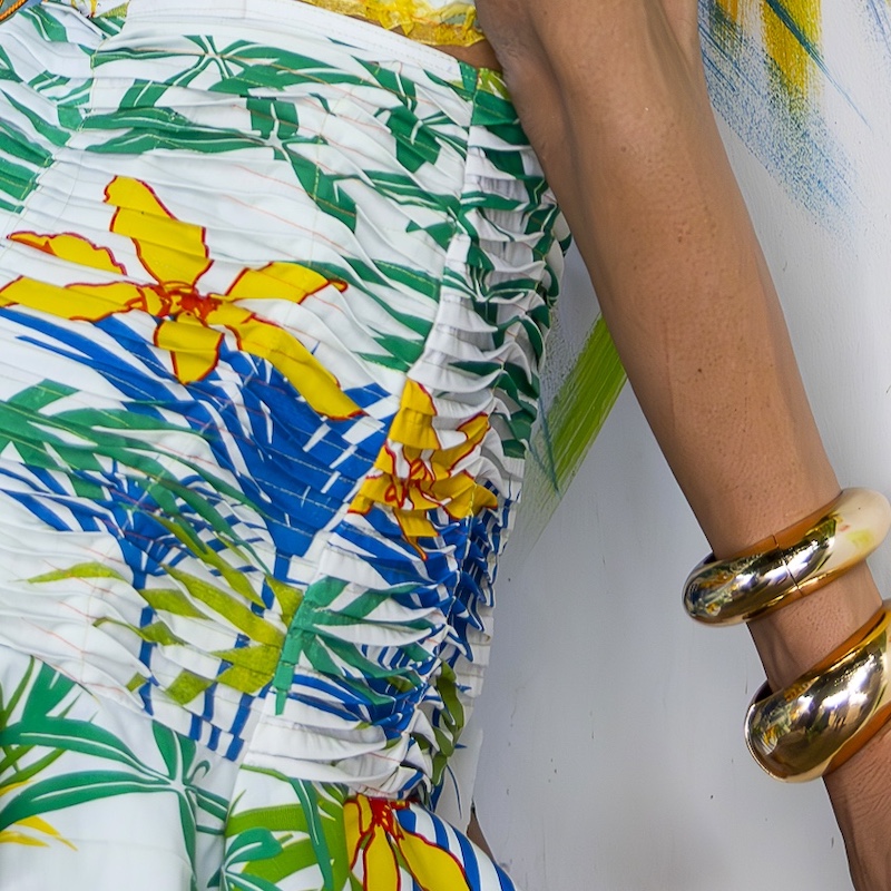 Uma pessoa vestindo uma saia branca com um padrão colorido de flores e folhas tropicais, em pé ao lado de uma parede com listras de tinta verde e amarela. A pessoa tem pulseiras douradas em seu pulso.