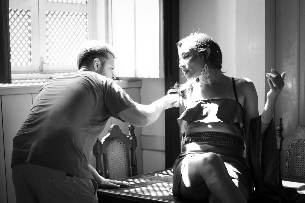 Uma pessoa ajusta a roupa de uma modelo sentada em uma sala iluminada pelo sol. A modelo está sentada em uma mesa, usando um vestido com uma perna cruzada, e olha para o lado enquanto segura um cigarro.