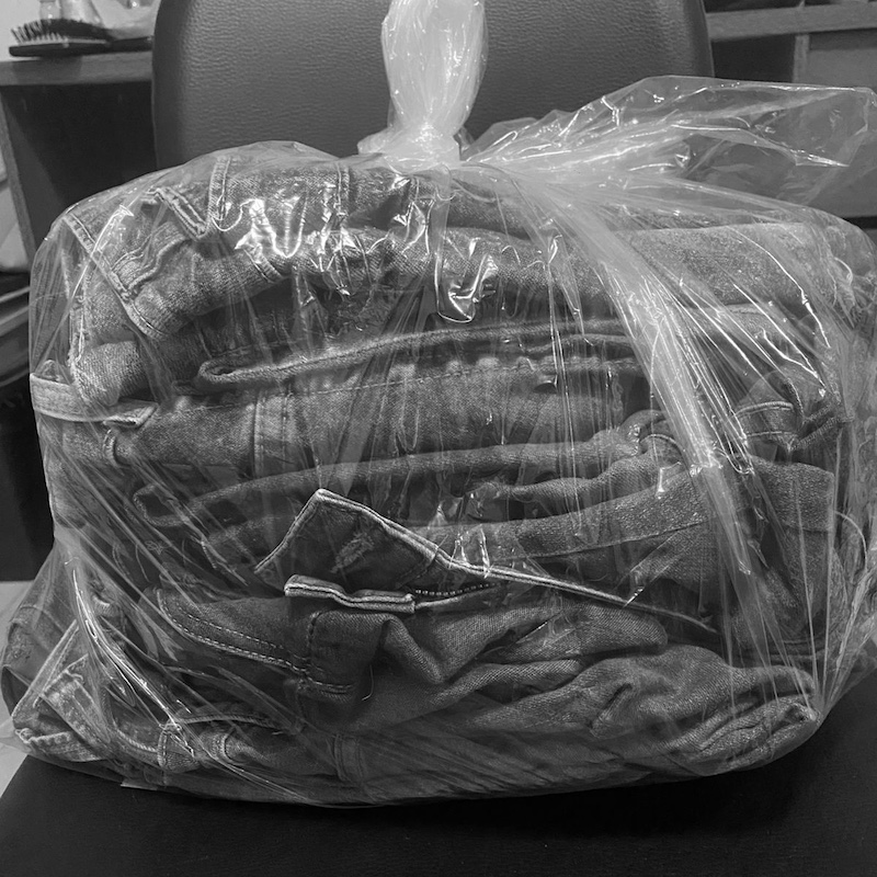 Um saco plástico transparente contendo uma pilha de jeans dobrados está sobre uma superfície escura, com uma cadeira visível ao fundo.